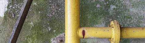 ...Lignes formées par une canalisation en partie rouillée et un bâton de bois sur un fond de vermoulu sur un mur d’une friche industrielle....